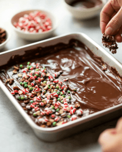 A baking sheet lined with parchment paper, covered with melted chocolate, as a hand sprinkles colorful toppings like candy canes and nuts over it.